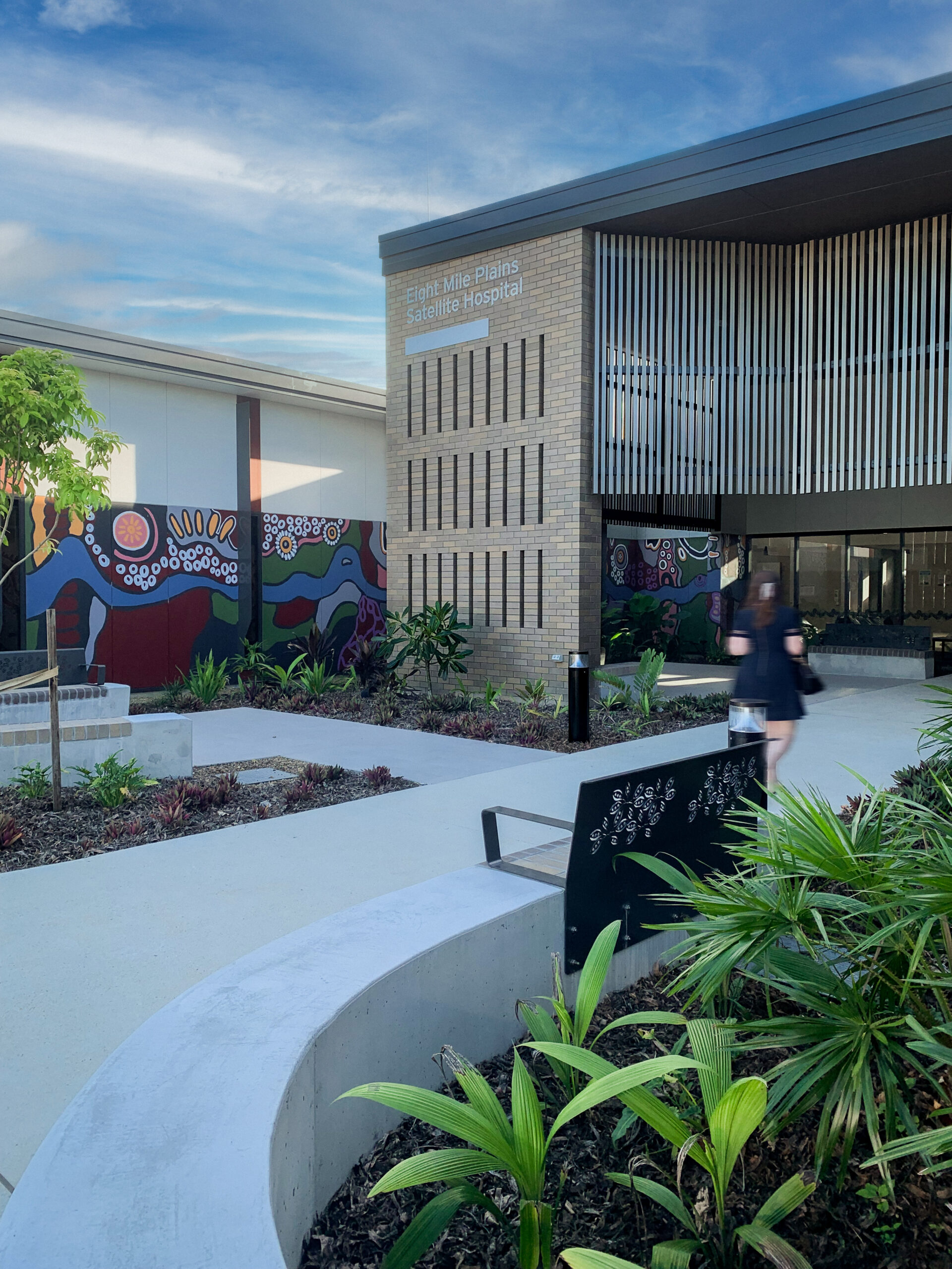 Female walking on a path towards the newly completed Eight Mile Plains Satellite Hospital, with green gardens and a Frist Nations artwork by Ailsa Walsh is painted on a wall mural.